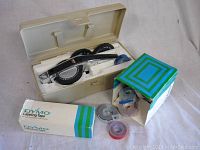 Photo showing the vintage Dymo label maker inside a beige plastic carrying case with two dials and the stamping lever visible, along with a couple of tape rolls outside the case.