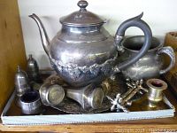 Photo showing the lot of vintage silver-plated items on a tray, including teapot, creamer, salt and pepper shakers, candle holder, small cup, and a decorative cross-shaped item.