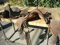 Two worn old western leather saddles on table outdoors, both with wooden horns and leather fringe.