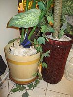 Two faux plants with one in a yellow-beige round pot and the other in a tall dark brown woven planter, with decorative shells visible on soil.