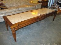 Full view of the long wooden coffee table with cream colored stone top on carpeted floor by a sofa.