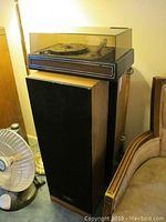 Photo showing the pair of Optimus speakers with wood veneer and black cloth covers. Turntable with clear dust cover placed on top of one speaker.