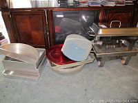 Overview of all items including warming pans, cake pans, plastic carrier, and chargers stacked on a carpeted floor in front of a wood cabinet
