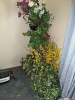 Metal black plant stand with multiple tiers holding faux plants and flowers including purple, white, red, yellow blooms and variegated green leaves in pots beside window.