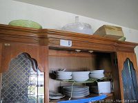Side view of china plates and bowls stacked inside wooden cabinet with glass doors, also showing items stored above the cabinet including green glass bowls and covered dish.