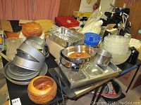 Wide view showing assortment of canisters, metal cake pans, plastic/acrylic bowls, wooden canisters, and kitchen utensils on folding tables.
