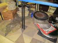 View of assorted cake supports, serving trays, and stacked platters with cake supports visible on the floor along with boxes and trays nearby