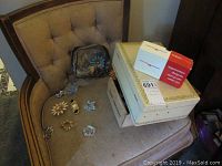 Assortment of costume jewelry and storage boxes arranged on a beige cushioned armchair, showing various brooches and vintage-style boxes.