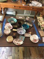 Overview of assorted home decor items including tea cups, pottery bowls, decorative plates, small boxes, and a glass jewelry box arranged on a table.