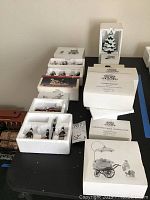 Overview of boxed and packaged collectible figurines and decorations from Department 56 Heritage Village collection on a black table.