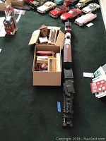 Full view of train set on carpet with boxes of assorted model train parts and vintage toy cars in background.