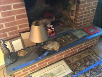 Two table lamps, one with beige shade and hand crafted metal base, second lamp black metal base; three picture frames of different sizes and styles; five pieces of hand painted tin art displayed on brick hearth.