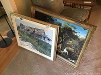 Two framed artworks leaning against a chair, one acrylic painting of a beach house and one print of Adam and Eve in a garden.