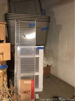 Four large grey plastic storage bins stacked, showing laminated surface and lids, with a white 3-drawer plastic storage unit beside them in basement setting.