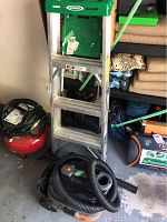Photo showing Werner ladder leaning in storage next to red and black Rigid wet dry vacuum with hose on garage floor.