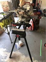 Full view of the entire compound slide miter saw mounted on the stand showing tripod legs and red adjustment knobs.