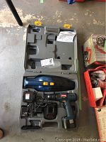 Gray plastic carrying case open showing Ryobi cordless drill driver, sub compact handheld vacuum, charger with cables, and instruction manual inside.