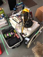 Overview of Wagner Power Roller Max paint sprayer with paint rollers and folded plastic tarps beneath, a bin nearby with boxes of nails and plastic tray.