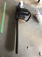 Full length top-down view of the Blue Max chainsaw showing the black chain guard and long cutting bar on a concrete floor.