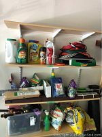 Three shelves holding assorted garden supplies: chemicals, seed mix, cedar shims, rubber garden borders, a plastic bin with tools and gloves, and various lawn care products in bags.