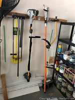 Four garden tools hanging on a wall: Fiskars lopper, Ryobi Expand-It trimmer attachment, spike aerator, and garden edger next to shelving with paints and supplies.