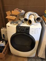 White front-loading gas dryer with black door, vent hoses and duct accessories on top, and boxes around.