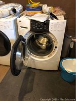 Front view of Kenmore Elite HE3T front-load washer with open door revealing stainless steel drum. Items surround washer including a blue bucket and trash can.