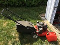 Side angle showing red Honda gas-powered lawn mower with black bag on green grass, near a garage door.