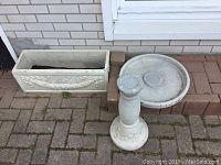 Photo showing a ceramic bird bath in two pieces, including a round basin and a pedestal, alongside a rectangular ceramic planter with floral relief designs, placed on an outdoor brick surface.
