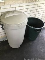 Photo shows two garbage bins side by side against a white brick wall. One lighter colored rubber bin features a lid and wheels. The other is open topped, dark green plastic bin without wheels.