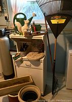 Photo showing garden tools including rakes, watering can, hand pruners, gardening gloves, gardening book and border decorations, along with garden pots and a small drawer unit.