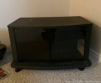 Front view of a black TV stand with two black-framed glass doors, set on carpeted floor against beige wall.