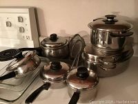 Photo showing stainless steel pots, pans with black handles and knobs along with a glass baking dish on a stove and countertop.