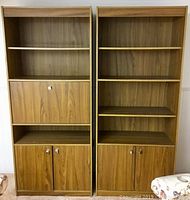 Two wood veneer bookshelves side by side showing shelves and storage cabinets, one with drop-down compartment and loose bottom piece.
