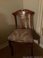 Front view of wooden chair with upholstered floral seat and carved backrest, shown in dim lighting near wall and curtain.