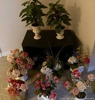 Nine flower pots arranged on floor and black cabinet, containing artificial roses, hydrangeas, and leafy greenery.