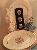 Photo showing three figurines including a white sculpted lady and a hand-painted lady in blue dress, a white decorative plate with floral pattern, and a black wall hanging with three oval scenic images.