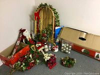 Photo shows an assortment of Christmas holiday decorations including a partially visible artificial Christmas tree, a wooden sled with some damage, multiple wreaths, a Santa figurine, floral sprays, boxes containing ornaments, and various other festive decor items scattered on the floor in a corner.