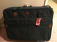 Full view of the green Jaguar suitcase with brown leather accents, front pockets, handles, shoulder strap and attached identification tag with red maple leaf.