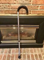 Photo of a height adjustable metal cane with a black curved handle and rubber tip at the bottom placed standing against a fireplace.