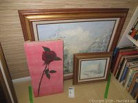 Three paintings leaning against wall: large landscape, smaller winter scene, and vertical rose painting on pink background