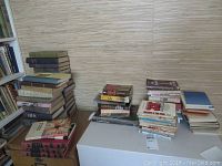 Overall view of three stacks of books, both hard and soft cover, placed on a box and table against a wall.
