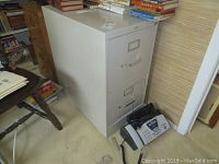 Two drawer metal filing cabinet in light tone with label holders and Brother fax machine on floor next to it, surrounded by books and furniture.