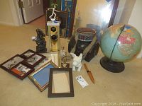 Photo showing all items in the lot arranged on the floor including globe, canteen, treasure box, trophy, dog planter, frames, bronze sculpture, and knife.