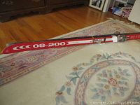 Photo of full length OS-200 ski showing red and white color, 79 inches long, lying on floor with carpet and wood background.