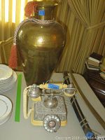 Photo showing large hammered metal vase with reddish tassel and rope by window curtains, alongside cream and silver vintage rotary phone on table.