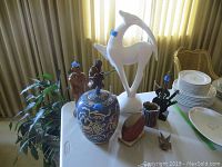 Photo showing full lot on table with potted plant near curtain, including two wood figurines, Asian vase with lid, white ceramic gazelle, metal guitar art, metal candle holder, some dishes partially visible