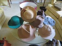 Overview of eight vintage hats on a glass table including the mink hat in box, green felt hat, straw hat, grey beret, and various brown/tan felt hats.