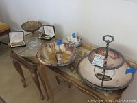 Silver plate serving bowl, brass compote, napkin holder, glass lazy Susan, and vintage cocktail forks arranged on a wooden table with floral detailing.