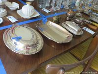 Photo of multiple silver-plated serving trays, dishes, and tableware arranged on a dining table, showing size and detailed edging on largest trays.